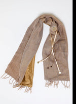 De duurzame, handgemaakte, betekenisvolle Plain sjaal in beige die elegant gedrapeerd is en een intrigerend patroon van verweven kleuren en texturen laat zien